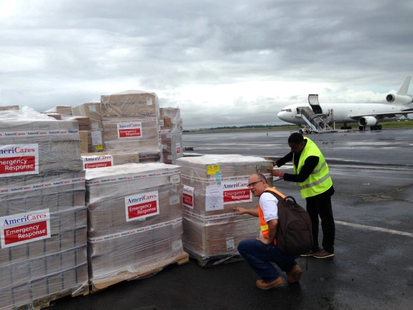 Americares supplies on shrink-wrapped pallets being checked by two workers on the tarmac with a supply plane in the background.